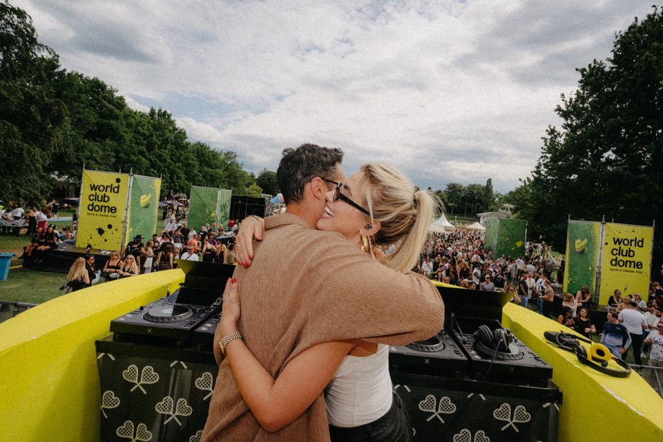 DJ und Freundin umarmen sich lachend am Ball-Pult, im Hintergrund die feiernde Wiese